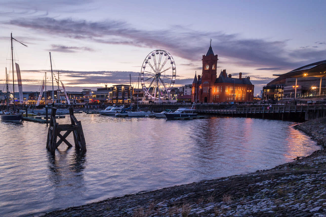 Twilight view of a waterfront featuring lit-up historical buildings, a ferris wheel, and moored boats with reflections in the water.